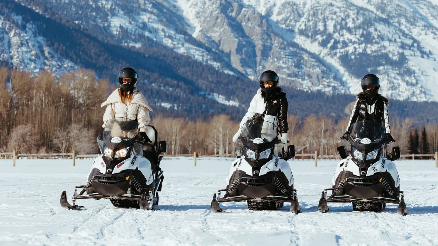 Three snowmobilers in stylish gear stand side-by-side on their machines facing the camera; a big snowy mountain looms behind.