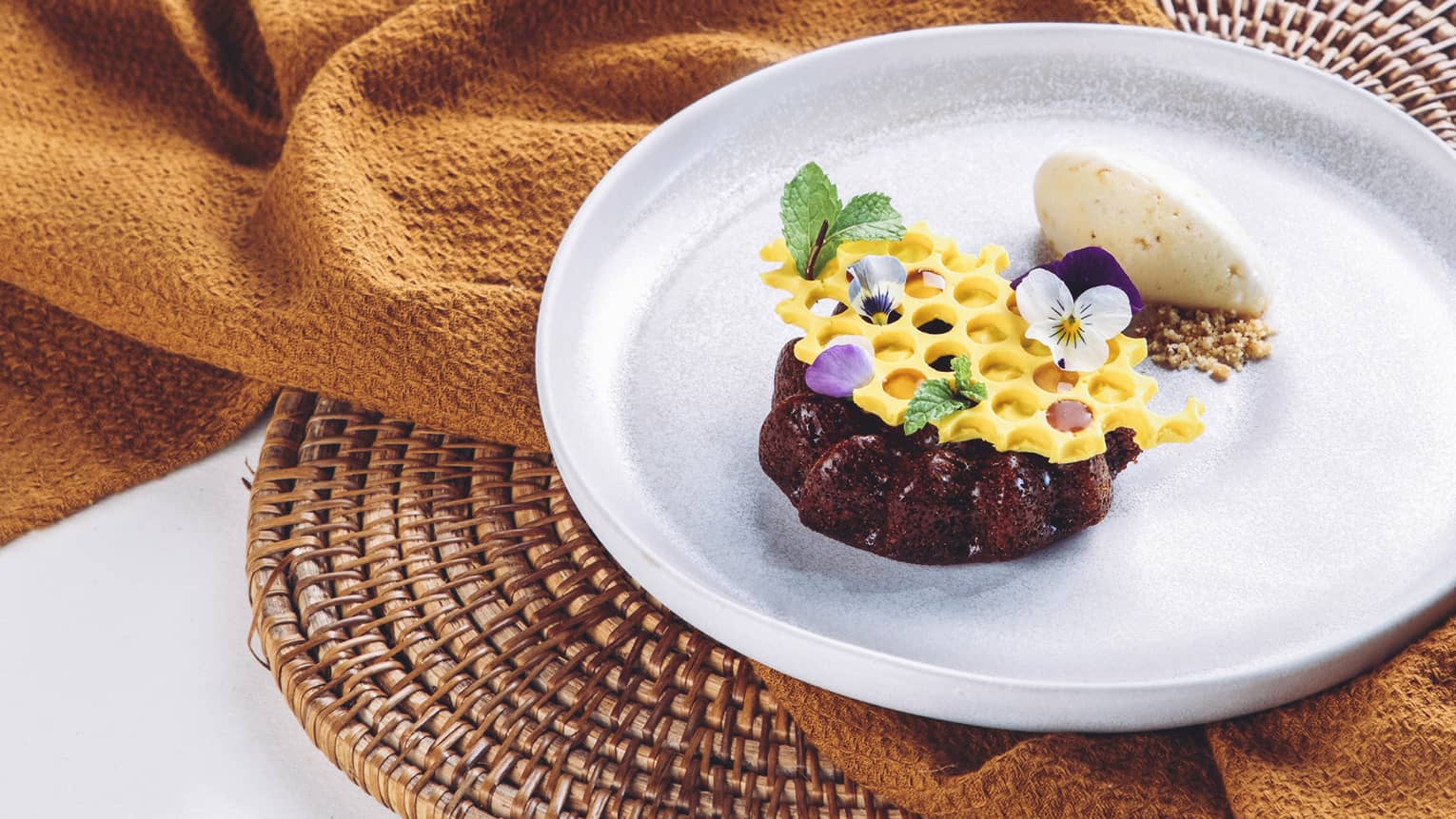 A rich chocolate mini-cake topped with honeycomb-shaped white chocolate garnished with mint leaves and dainty flower petals