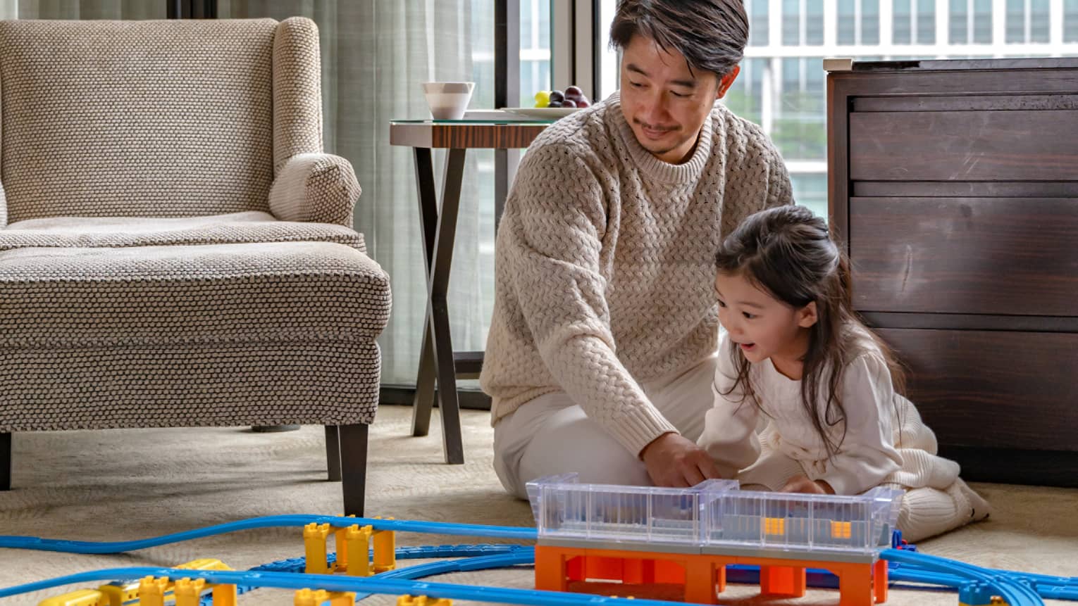 Man and young girl play with toy train set in guest room living area