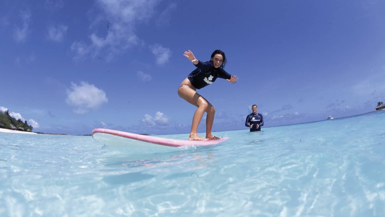 In the crystal-clear shimmering water is a woman learning to surf and an instructor standing in waist-deep water clapping.