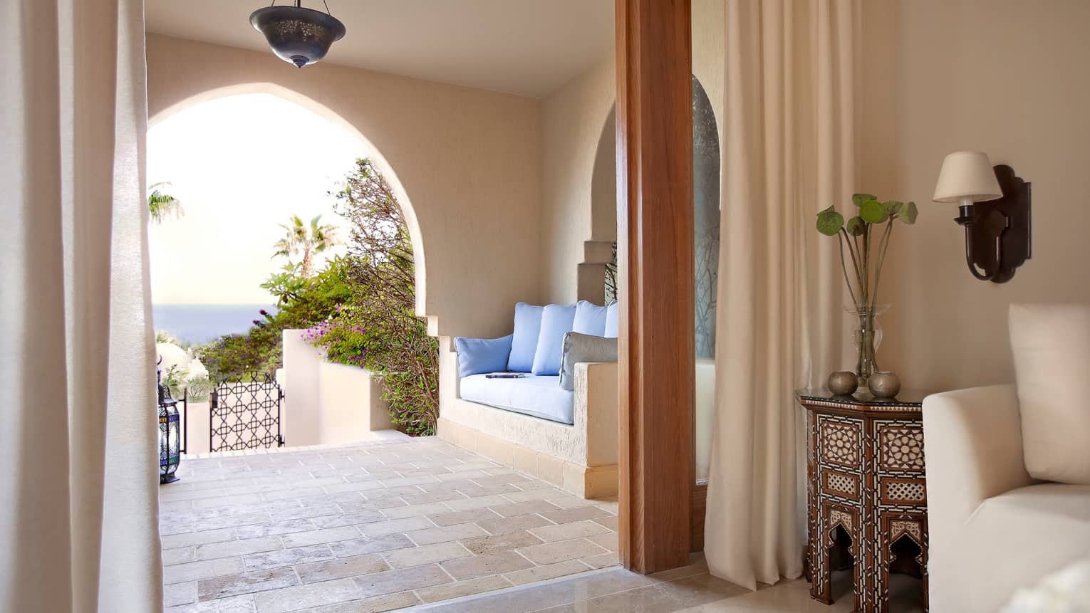 Covered balcony with cushions over clay sofa, tile floor, doorway to open-air room with decorative table, flowers
