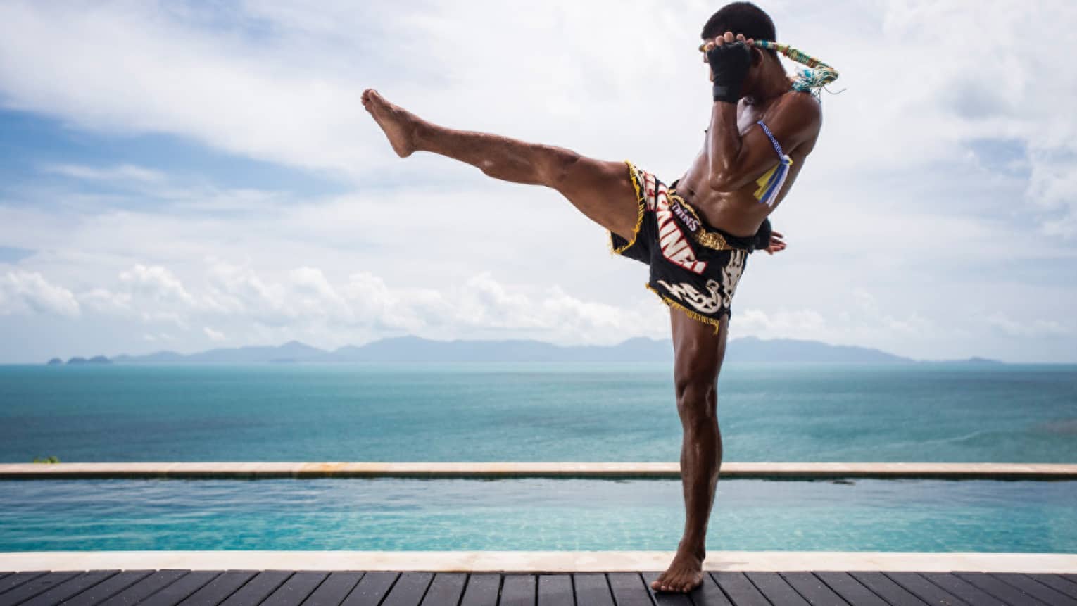 Side view of Muay Thai kick-boxer wearing shorts, doing leg kick on swimming pool deck in front of ocean