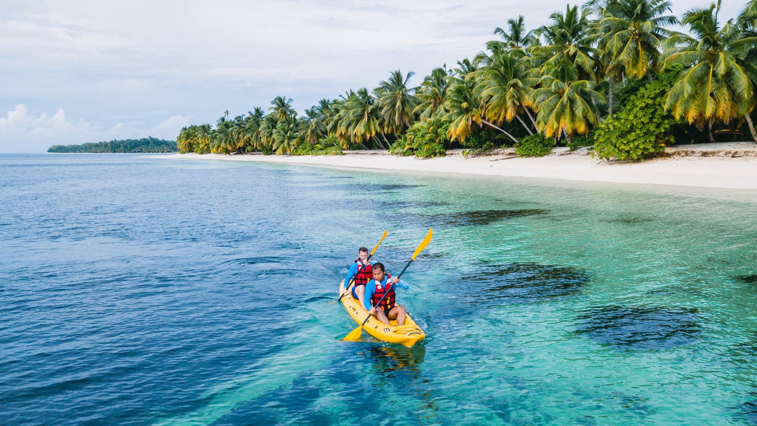 Two men wearing life jackets in yellow kayak paddle in ocean with palm trees in background