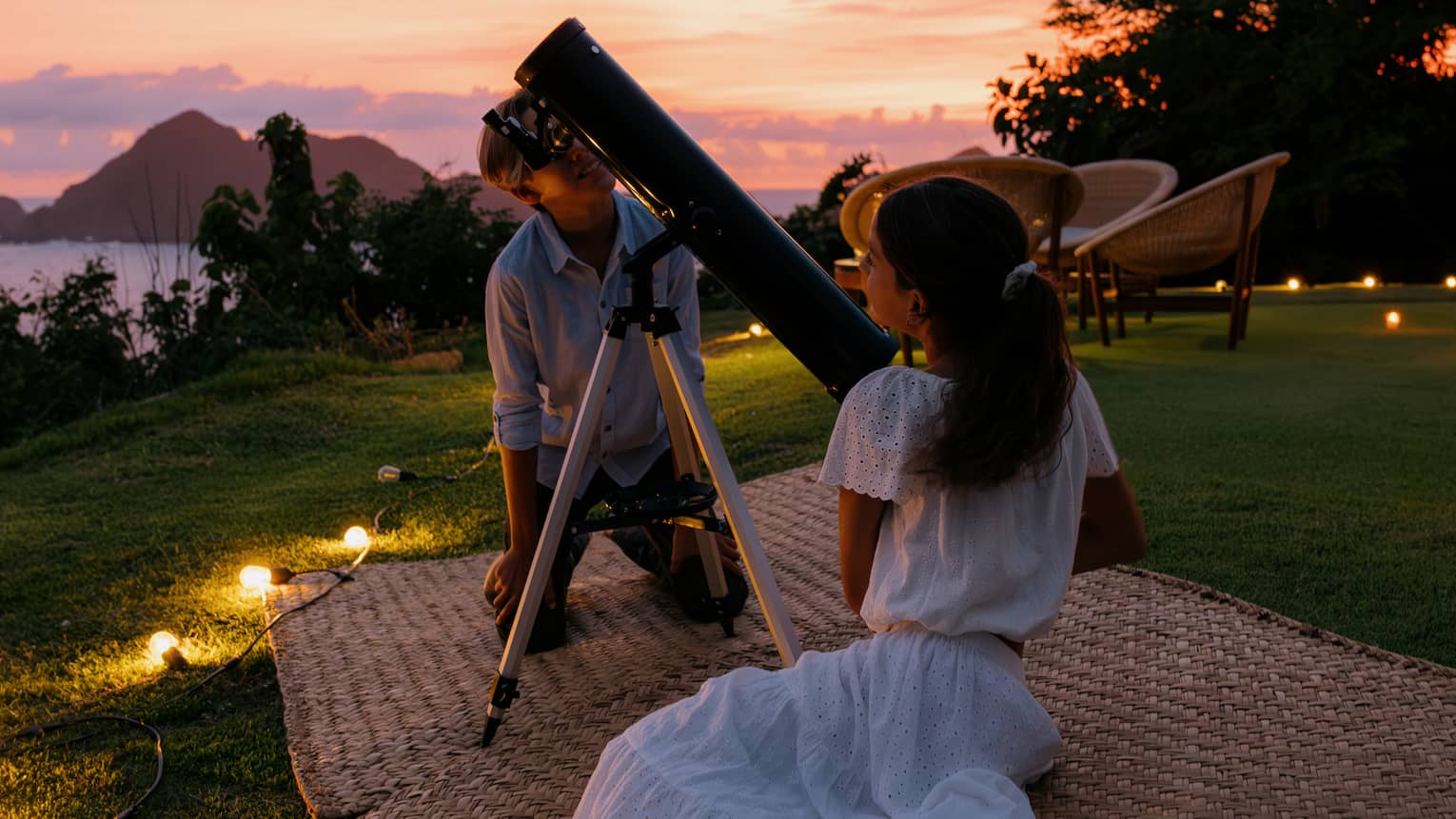 Two people on mats on the grass, with a large telescope. The ocean and a colorful sunset sky are in the background.