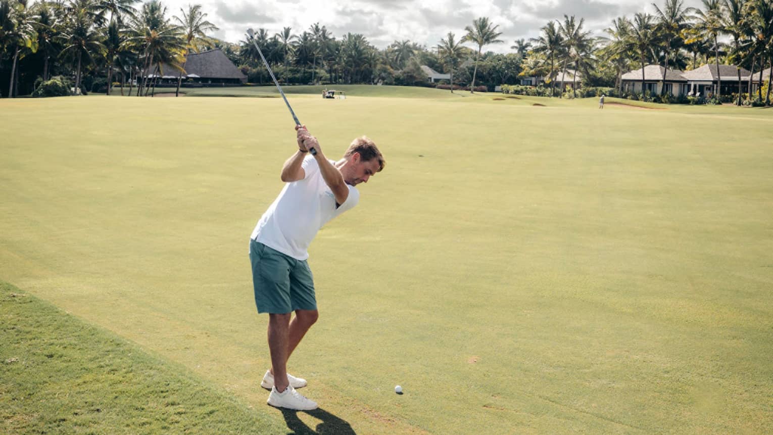 Man in white polo and green shorts swinging a golf club on green with palm trees and cloud-dotted sky in backdrop