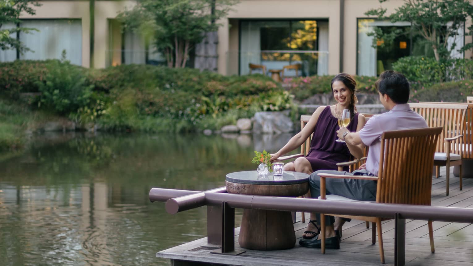 Smiling couple clink champagne flutes at a café table on a wood deck overlooking a rippling pond bordered by lush foliage.