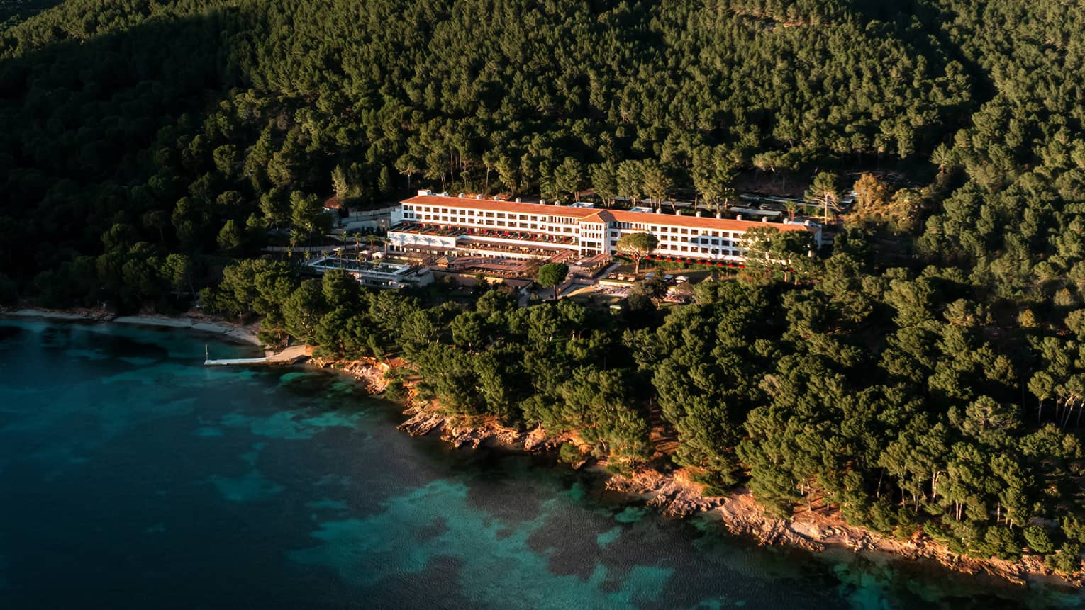 An aerial view of a wooded, mountainous coastline with a resort building among the trees, blue water in the foreground, and mountain views and blue sky in the background