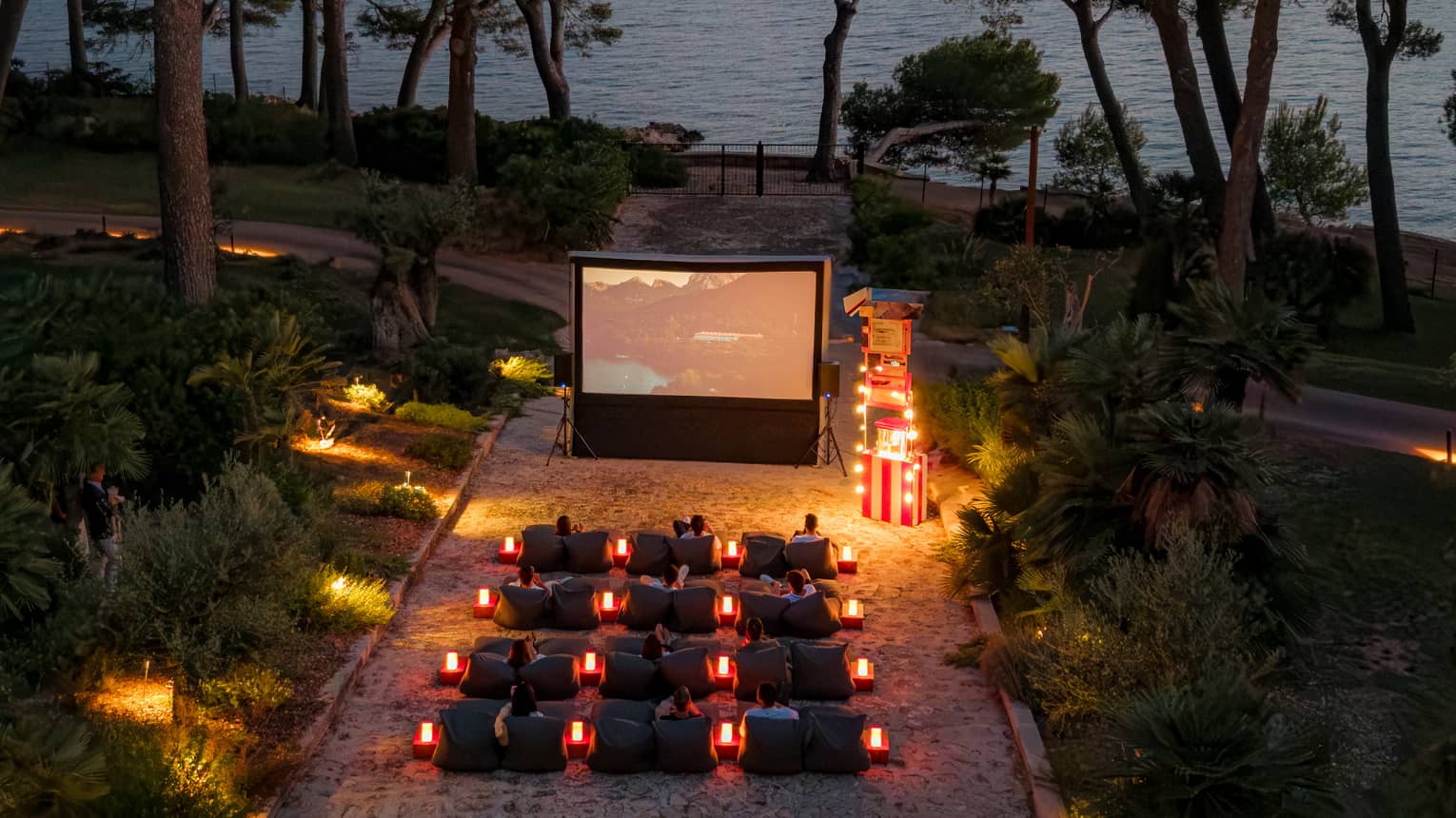 A bird’s-eye view of people seated in rows of chairs in front of a large movie screen in an outdoor setting in the evening, surrounded by trees, with the ocean in the background