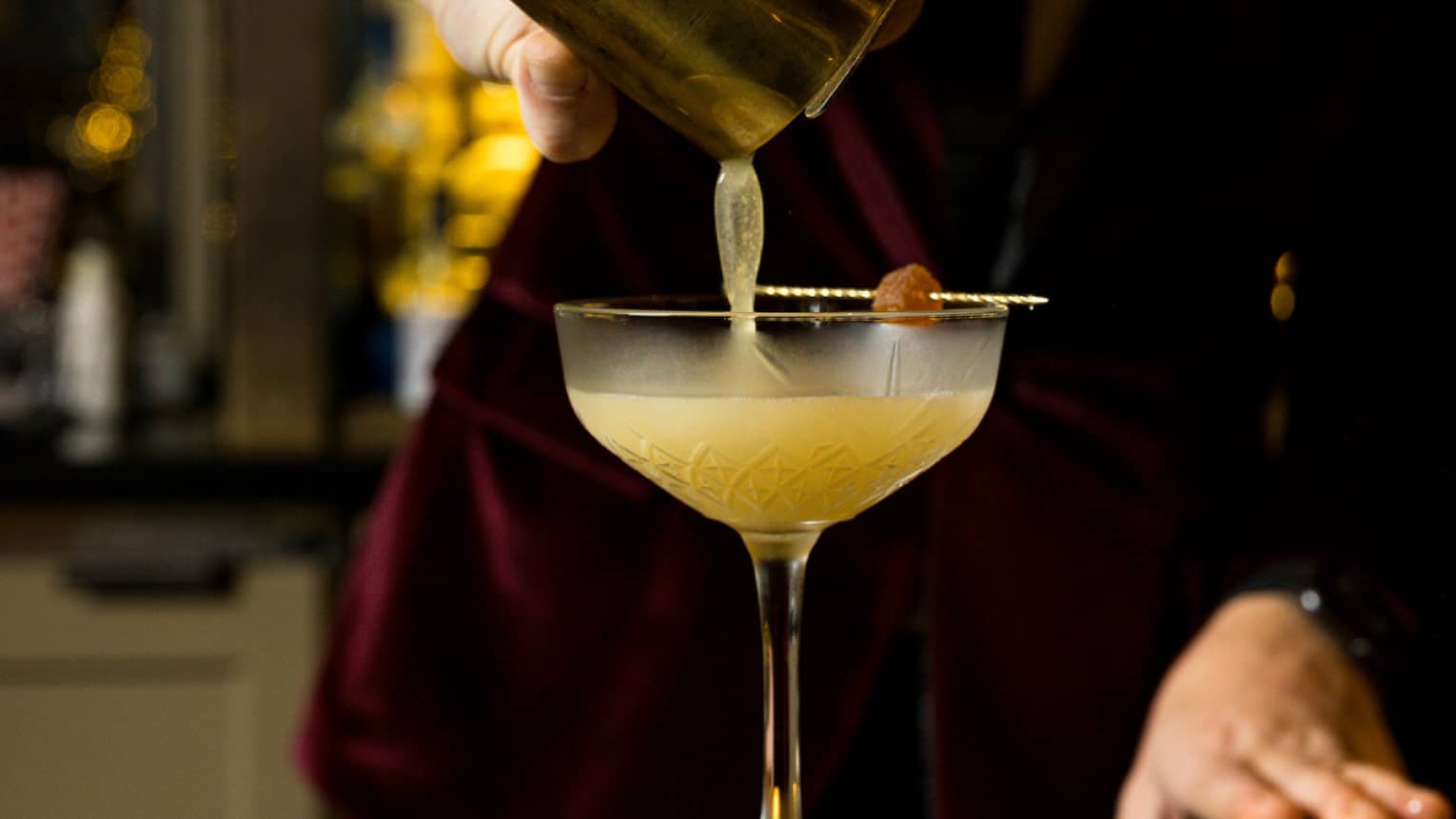 Bartender wearing a burgundy velvet suit and bow tie pours a cocktail from a shaker into a stemmed glass