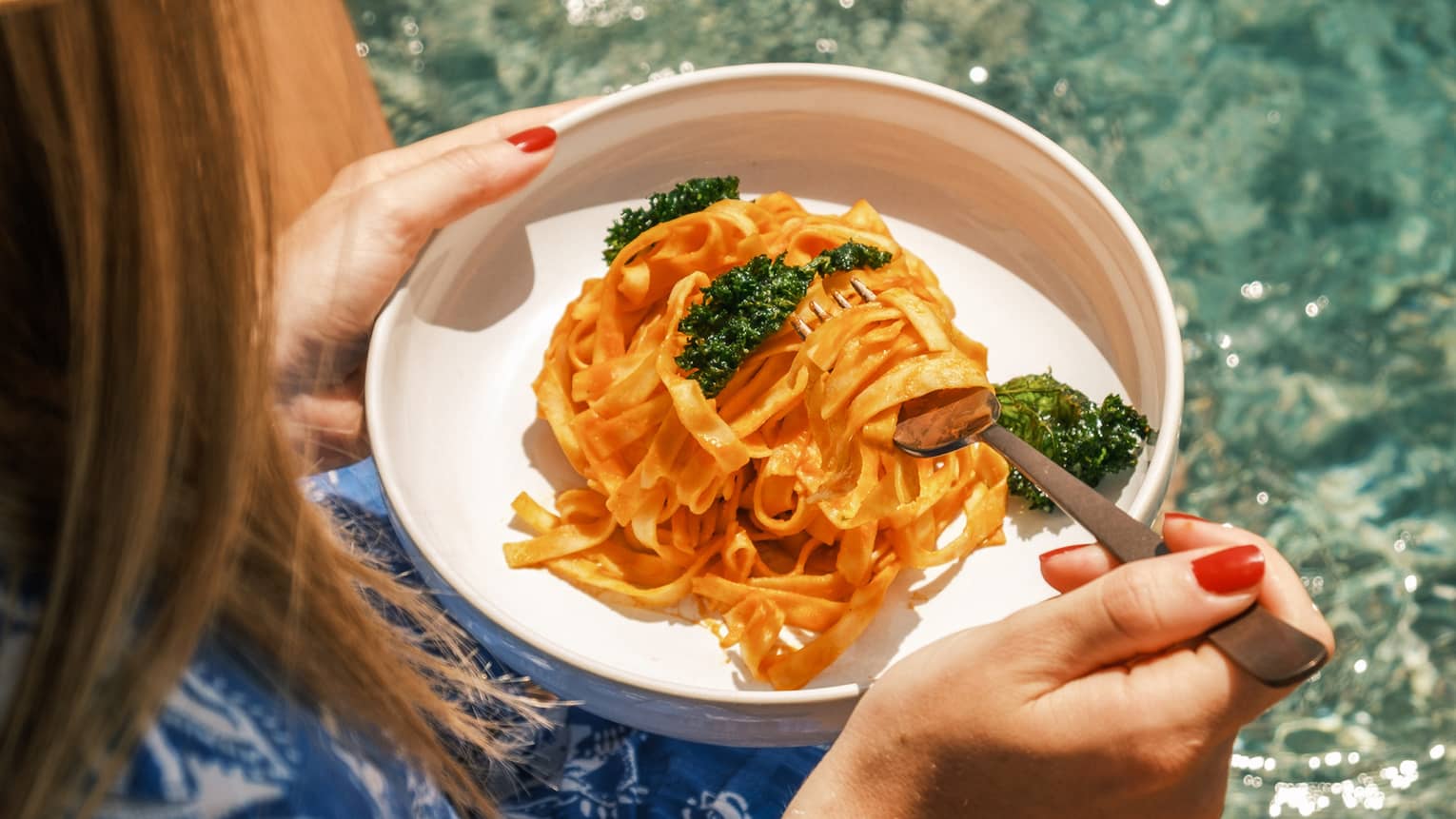 A guest wearing a sun hat and a blue and white dress sits on the edge of a pool and holds a plate of pasta and a fork.