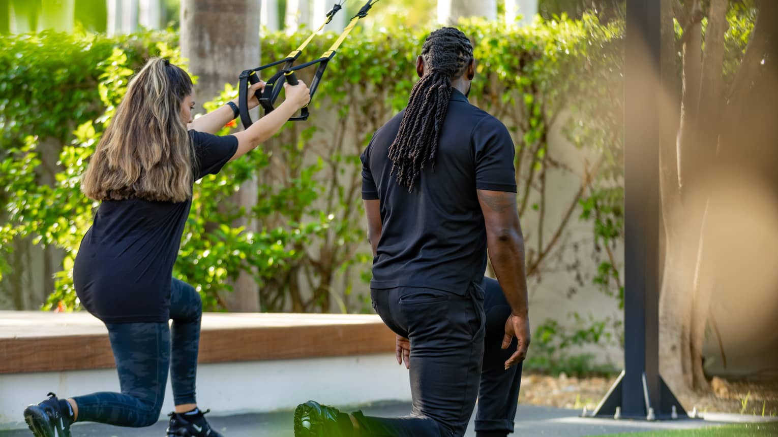 A guest practices lunging with suspended resistance bands in an outdoor, tree-shaded gym space. alongside a trainer.