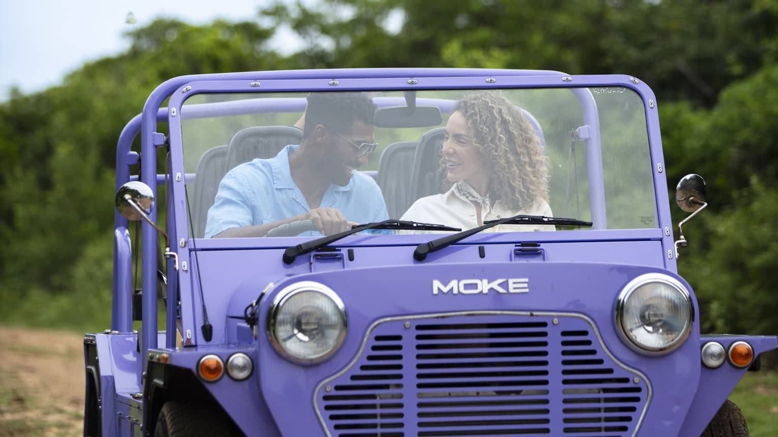 Front view of a couple driving a purple Moke, a small open-air recreational vehicle, along a dirt road surrounded by trees.