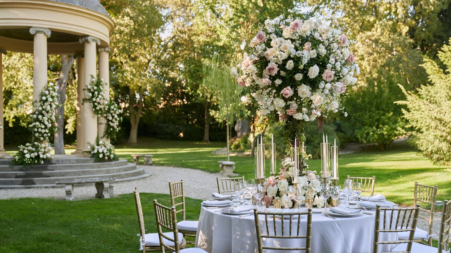 White formally set wedding reception table in garden with large white topiary and gazebo in backdrop