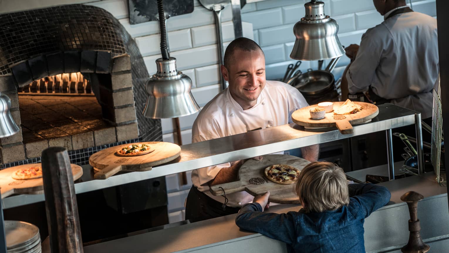Chef at open kitchen line near wood-burning oven passes wood tray with small pizza to child