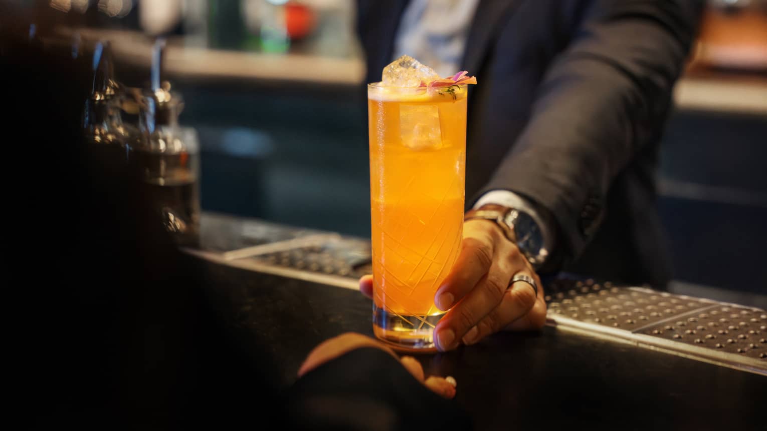 A person sets a bright orange drink in a tall clear glass on a bar counter.