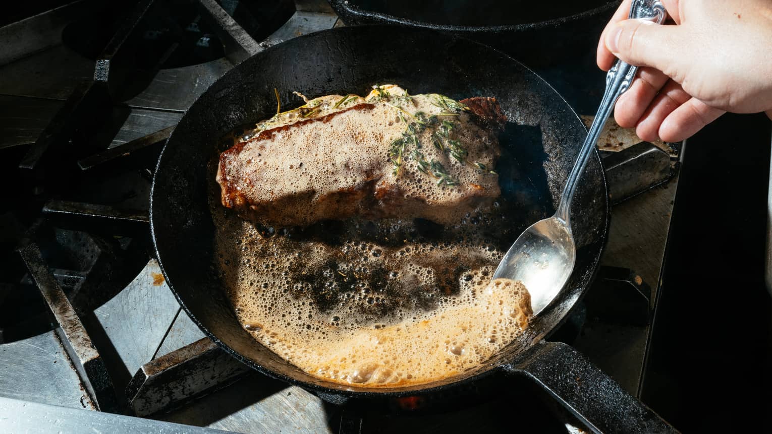 A skillet with a garnished cooked cut of meat and a pool of bubbling sauce, which a chef is scooping with a spoon