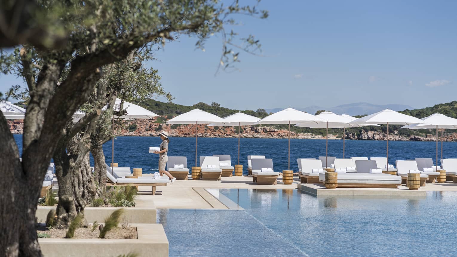 Woman holding towel walks past hotel pool with white chairs and umbrellas beside blue ocean
