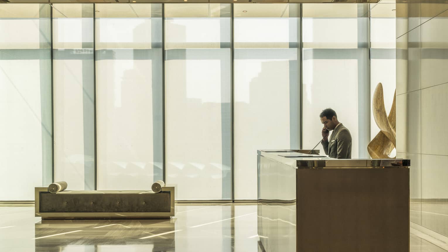 Hotel staff answers phone at modern reception desk near wall of windows, chaise on marble lobby floor