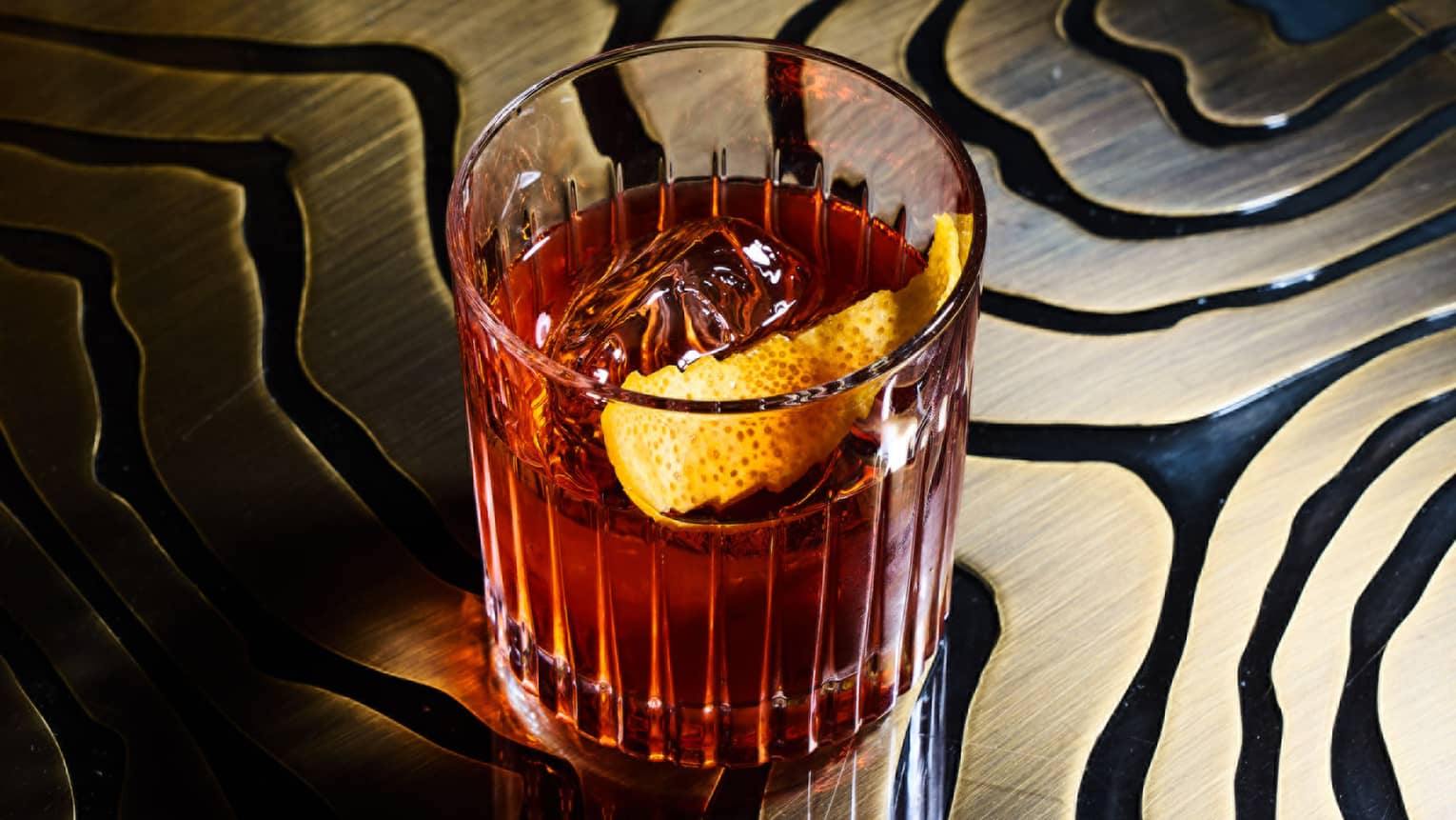 Old Fashioned cocktail with ice and orange rind in a clear Old Fashioned glass, sitting on a black and gold table
