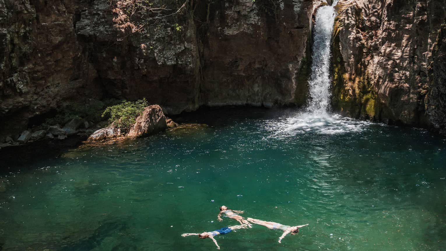 Three people float in a lagoon near a waterfall