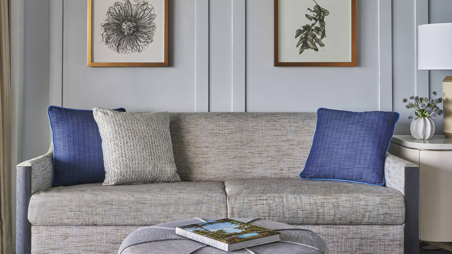 Two-seat sofa and grey, round ottoman/table, set against a wall with four hanging flower pictures