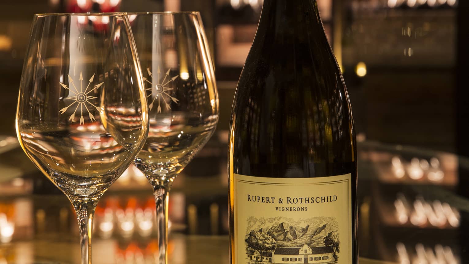 A close-up of two sparkling wineglasses and a dark bottle of sealed Rupert & Rothschild wine.