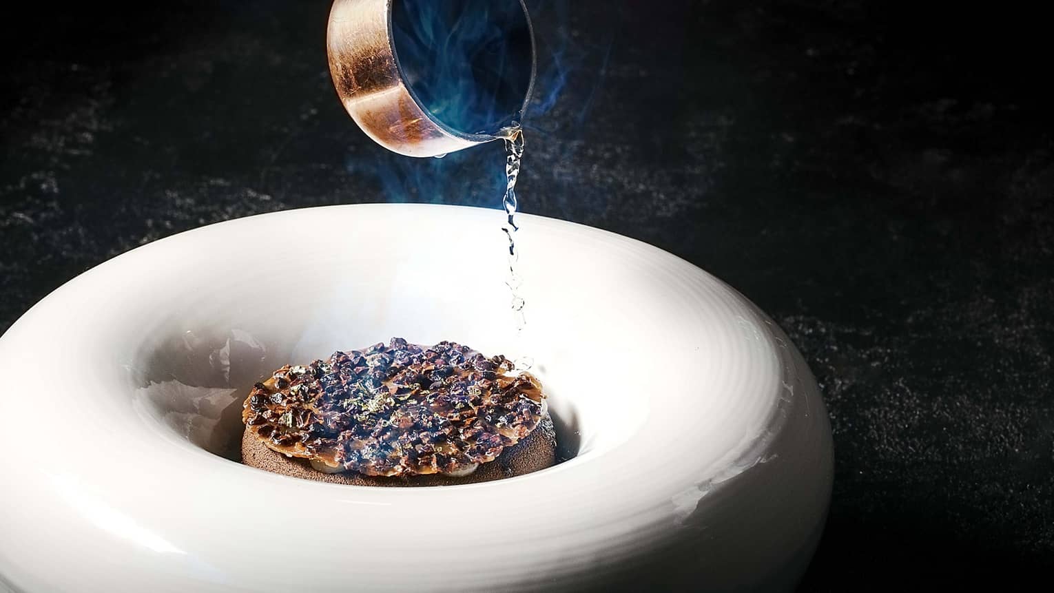 Sauce from a mini copper pot is poured onto food served in a large donut-shaped white bowl