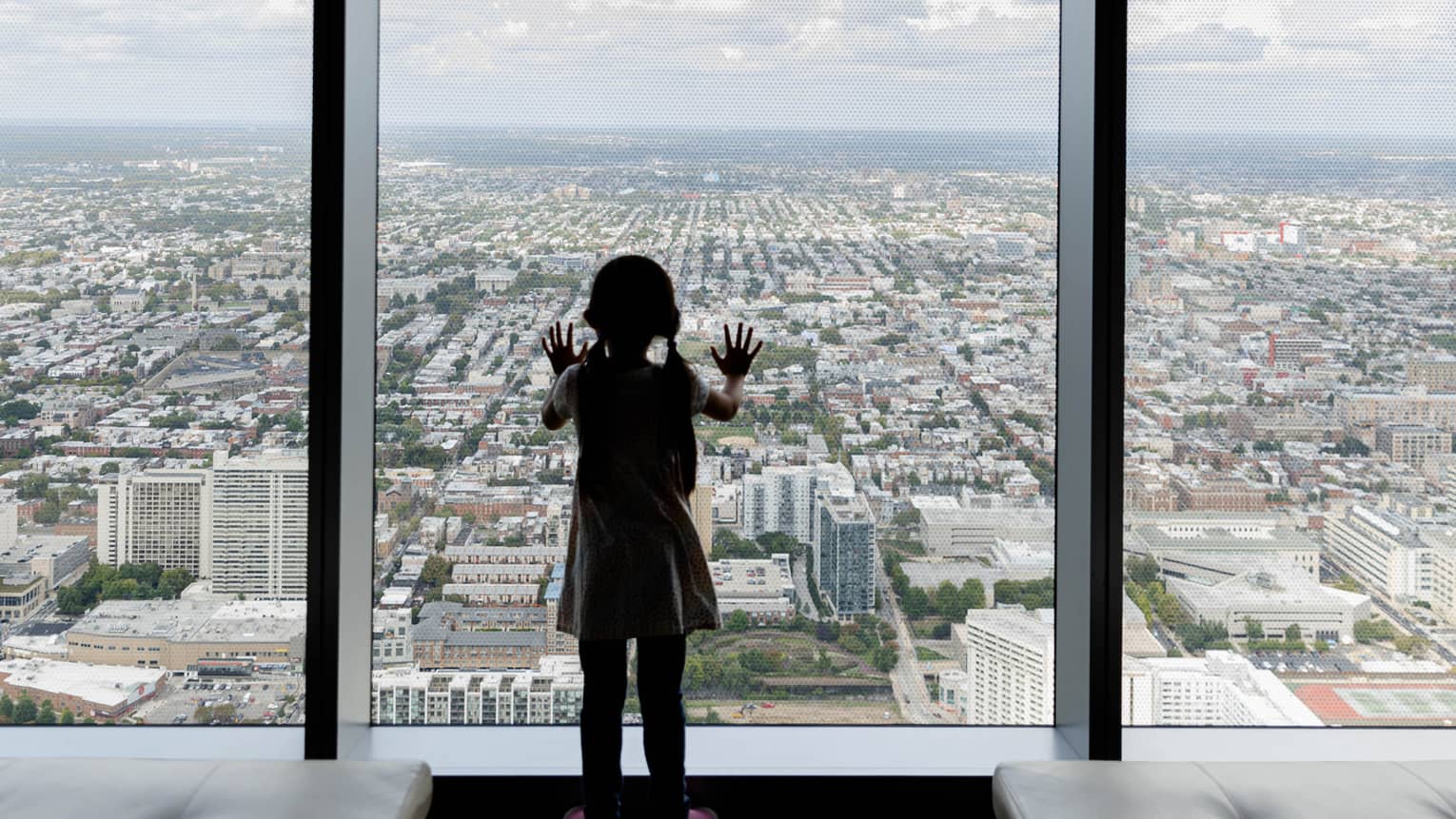 A young girl looking out of a large window in a tall building at a city below.