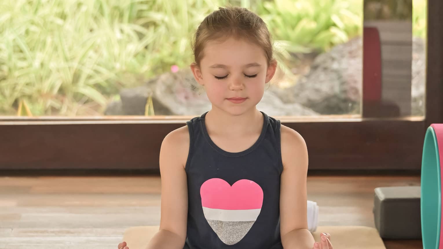 Child sitting in crossed-legged yoga pose on beige mat with eyes closed
