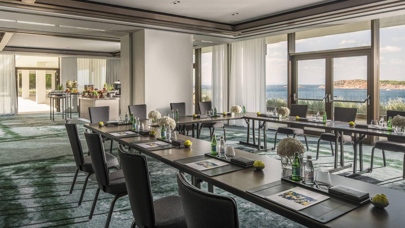 Sunny meeting room with tables in square, water, flowers and buffet, overlooking ocean