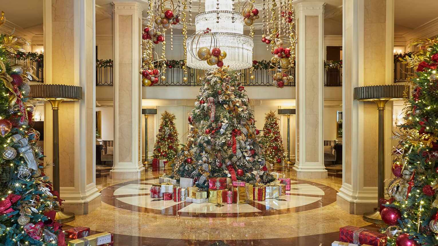 Red and gold decorated Christmas trees sit in the lobby under a tiered chandelier as ornaments handing from the ceiling
