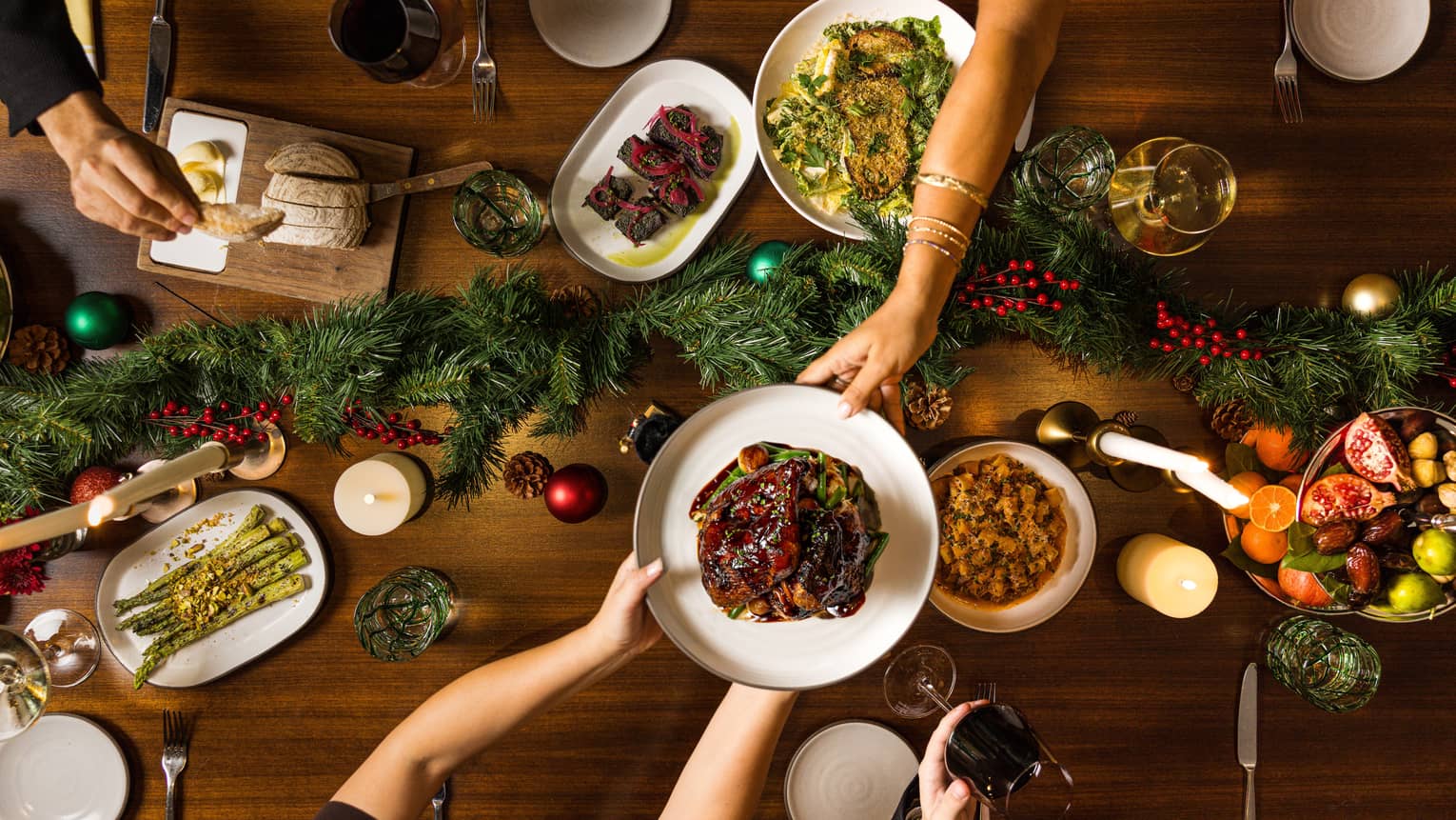 Hands pass a dish of food to another over a table set for a holiday meal with a green garland in the centre