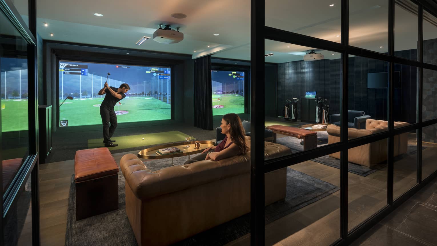Topgolf lounge, man swings club by large golf simulator screen, woman sits on leather sofa behind him