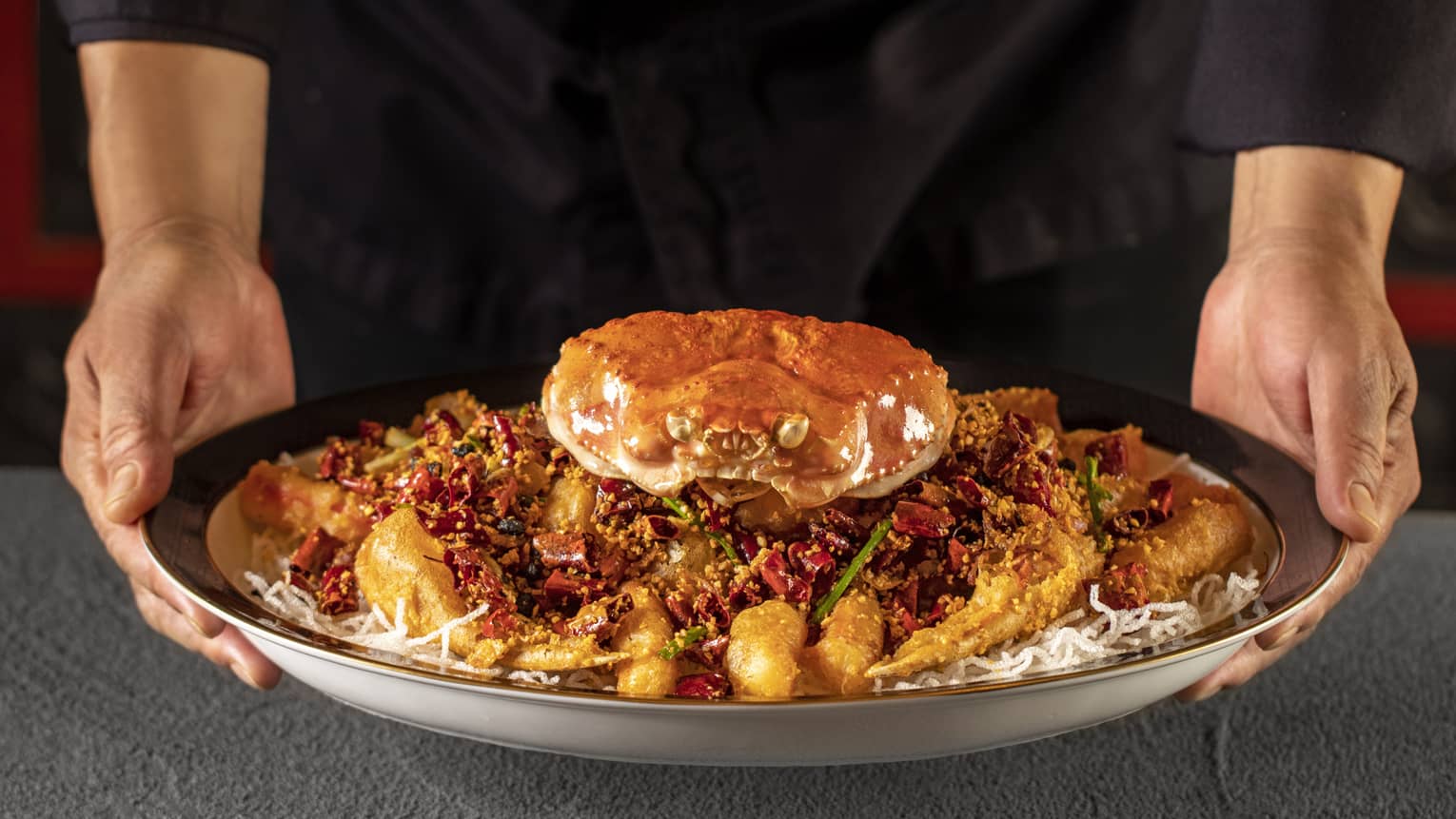 Two hands hold dish of whole crab on bed of crispy rice noodles