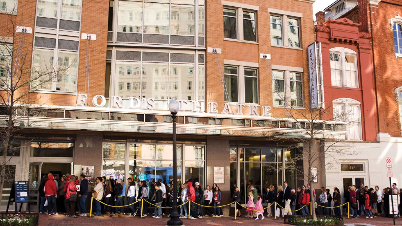 People lined up along sidewalk under brick building, awning with Ford's Theater sign
