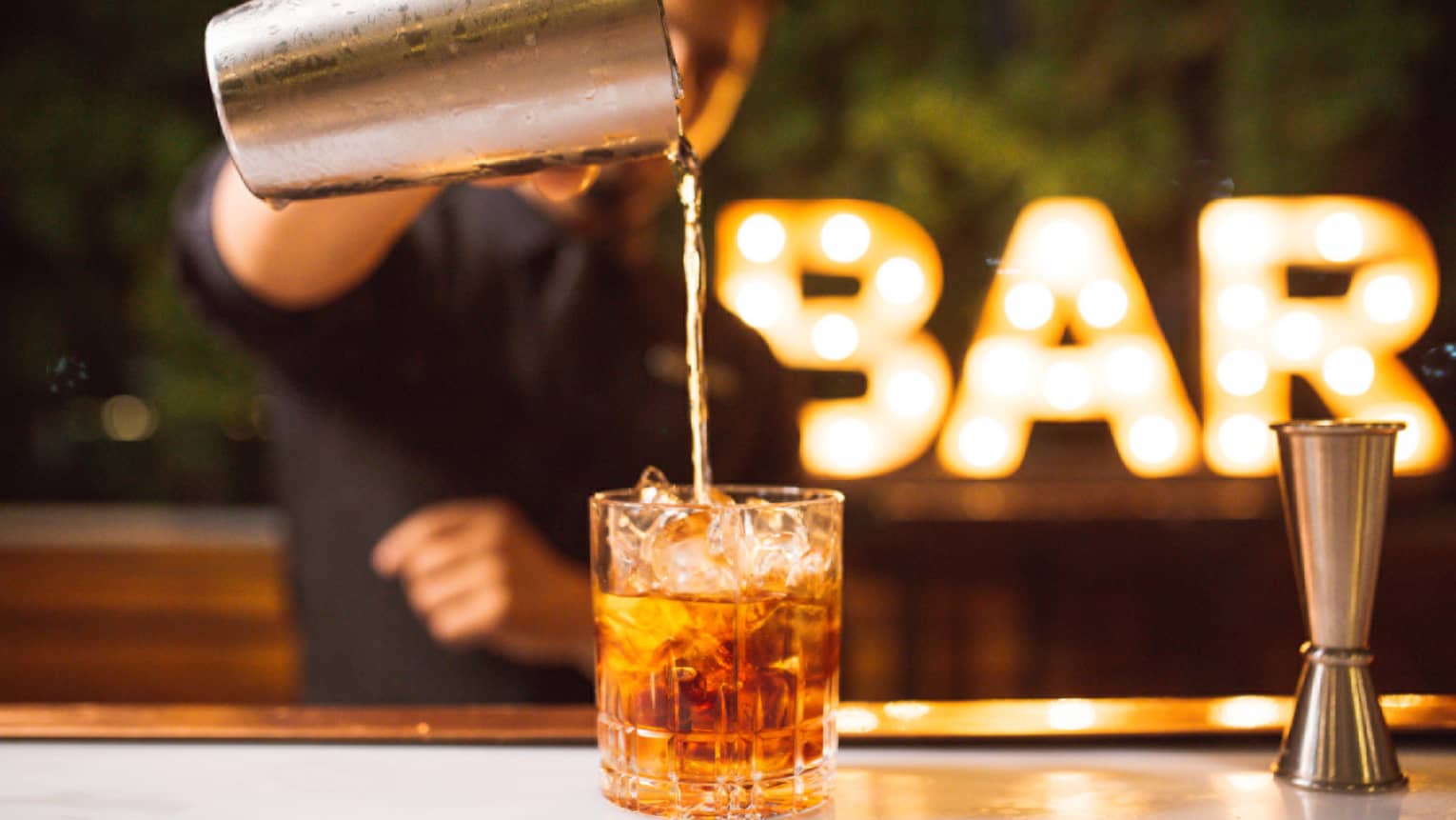 A bartender pours a glass of brown liquor with an illuminated Bar sign in the background