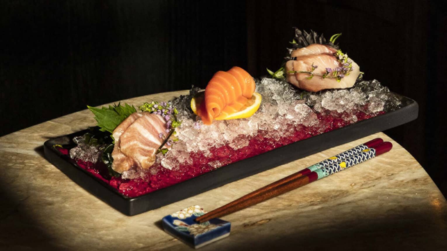 A plate with slices pink- and orange-fleshed fish sits on a table next to a set of chopsticks.