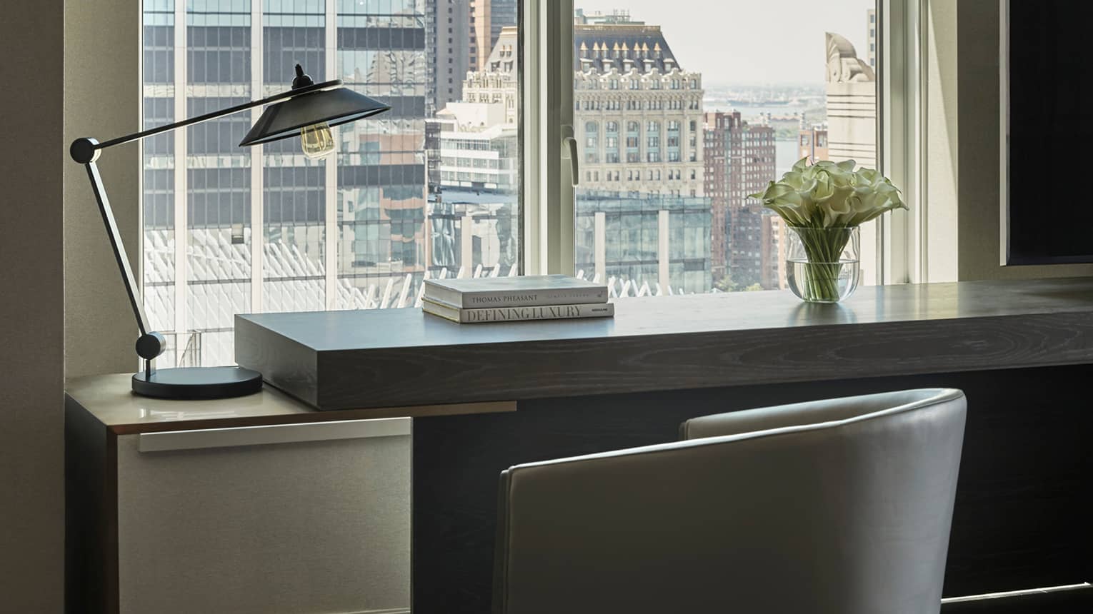 Hotel console table with a modern black lamp, a stack of books and a vase of white flowers, set against a window overlooking city buildings