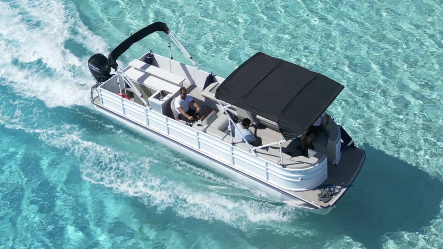 Aerial view of a pontoon boat in pale turquoise waters; guests are relaxing beneath a small canopy at the bow.