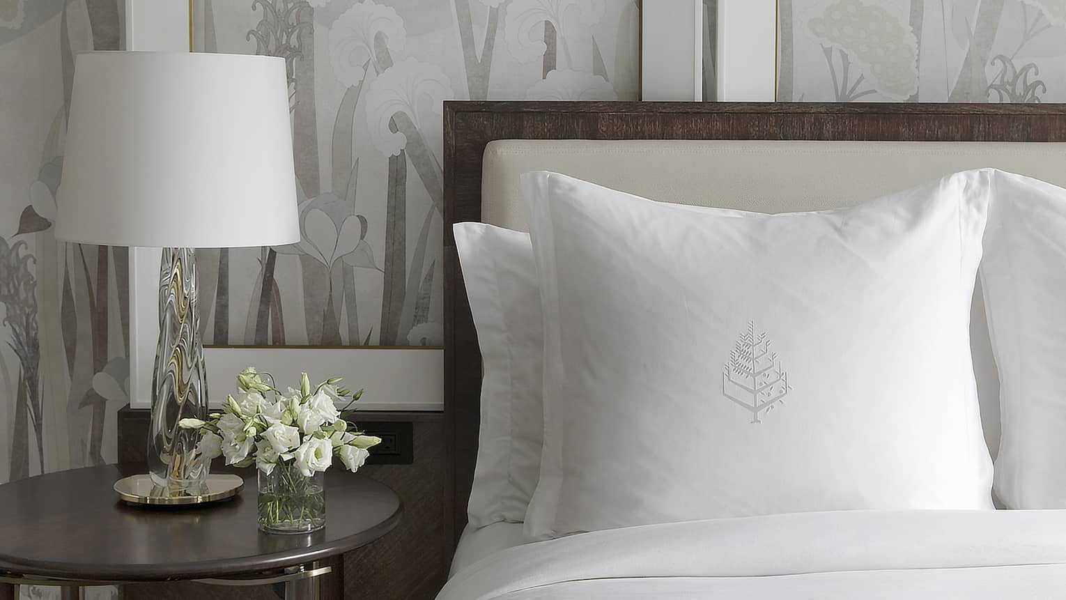 White flowers decorate a bedside table next to a bed with crisp, white sheets