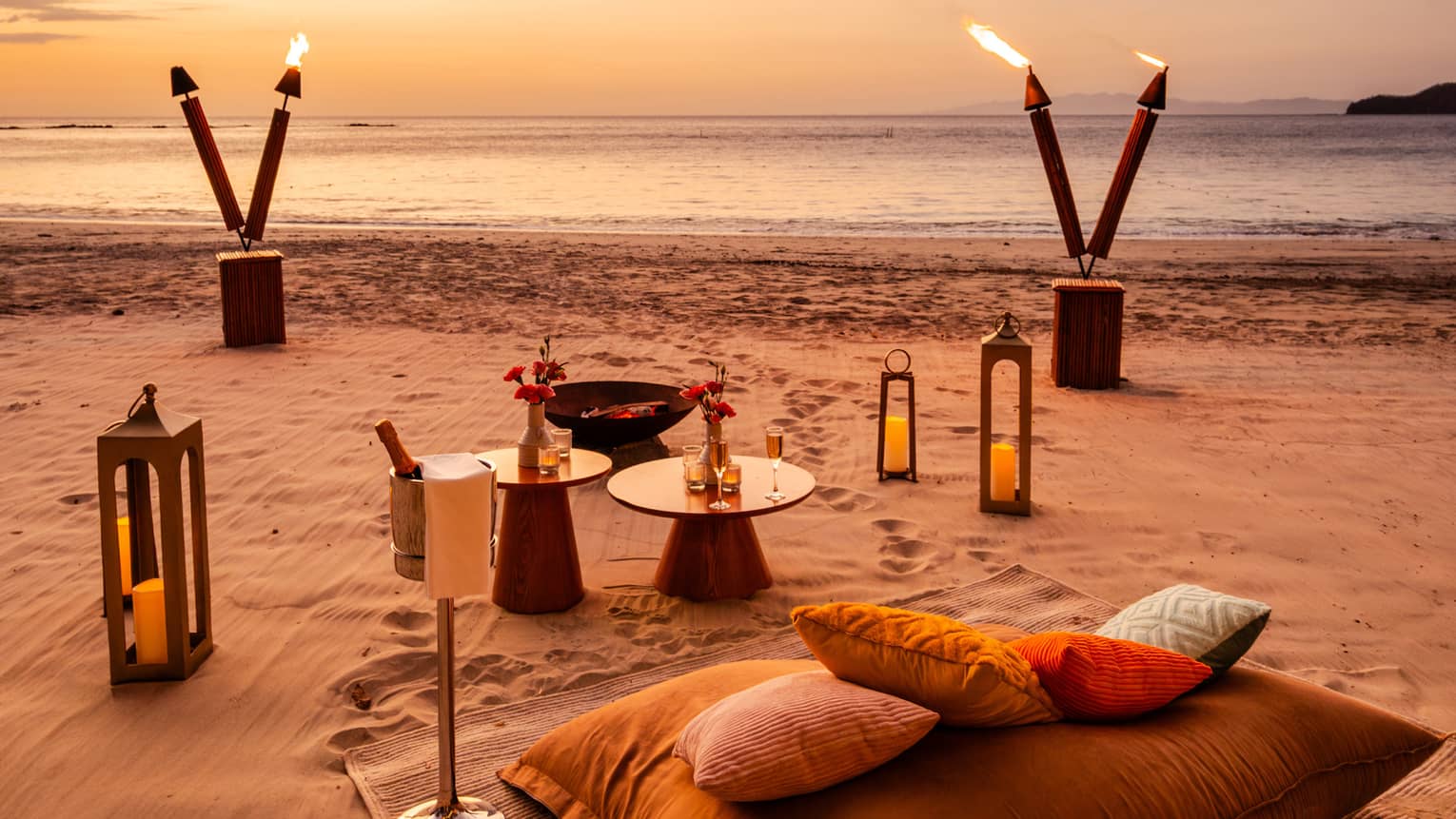 Blanket, cushions, small tables, lanterns and tiki torches are set up for a private dinner on a beach at sunset
