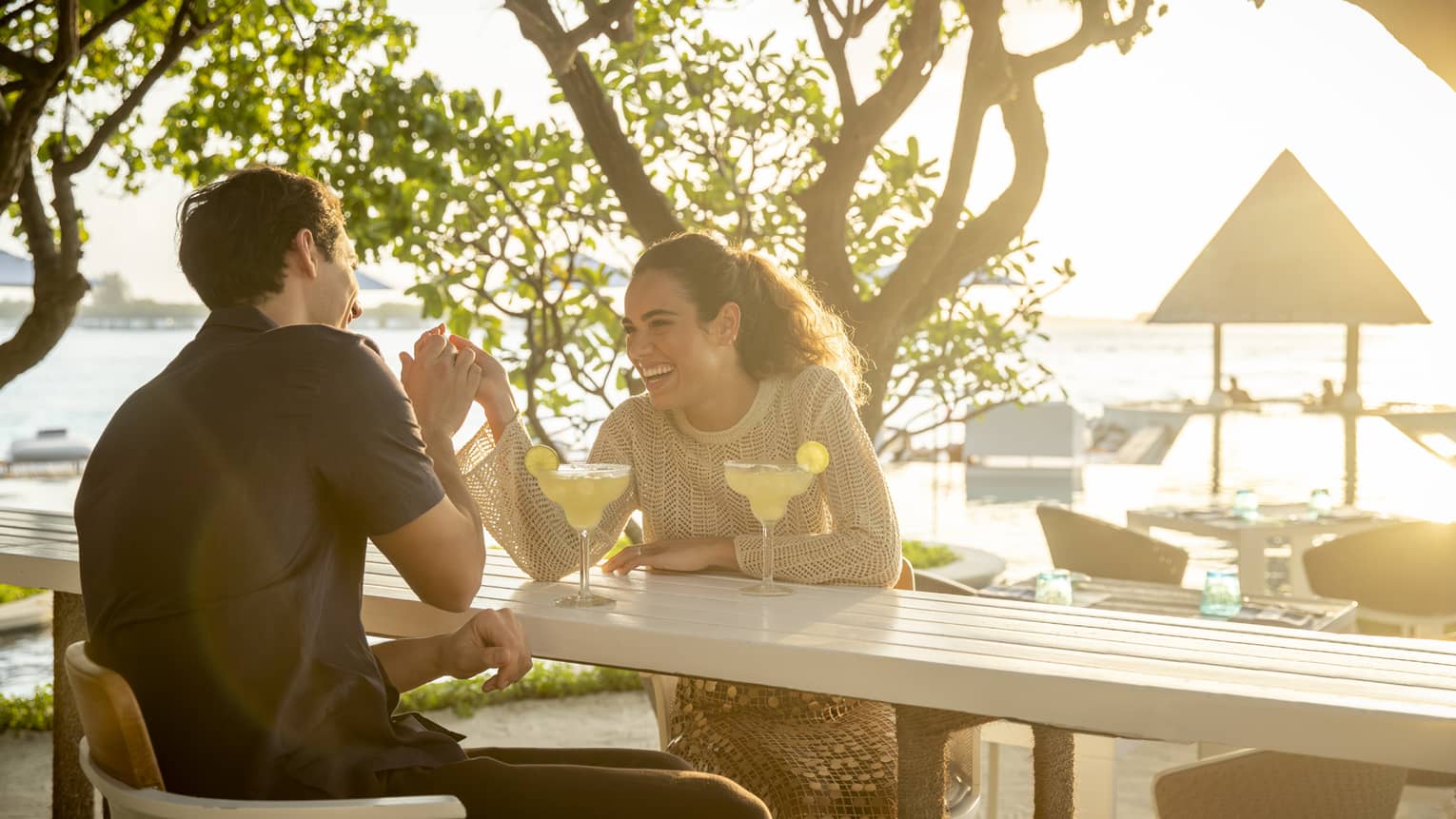 Laughing couple enjoys cocktails at outdoor beachside restaurant surrounded by greenery