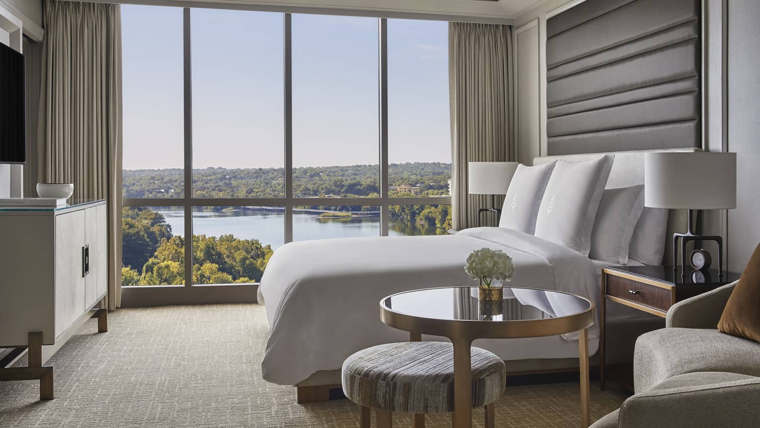Side view of small brass and glass table, hotel bed by floor to ceiling window and lake views