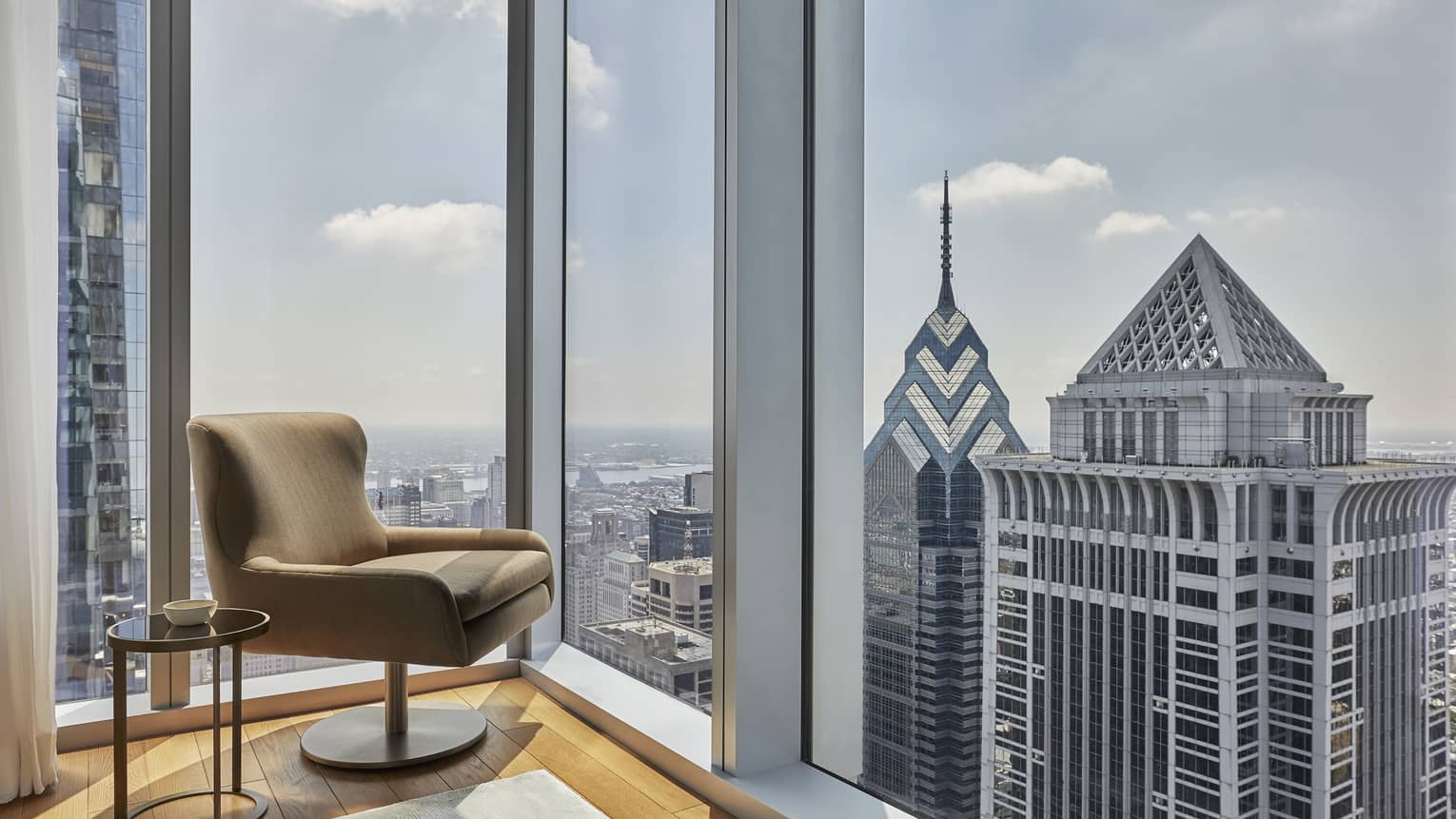 The corner of a hotel suite with large windows and a chair.