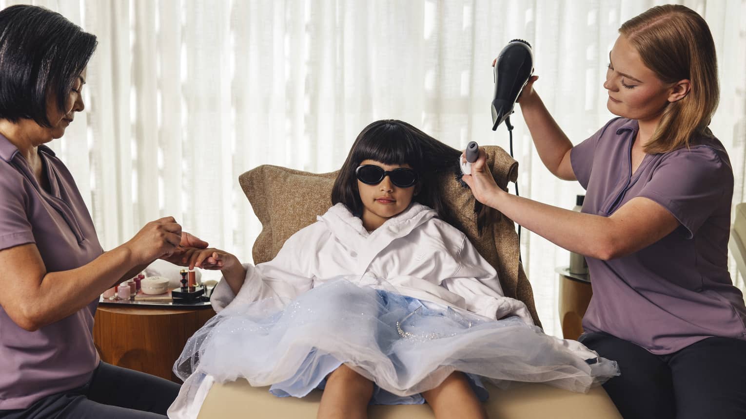 Little girl with dark brown hair and bangs lounges on a beige seat wearing a light-blue princess dress, a white robe and sunglasses as one spa attendant blow dries her hair and another paints her nails