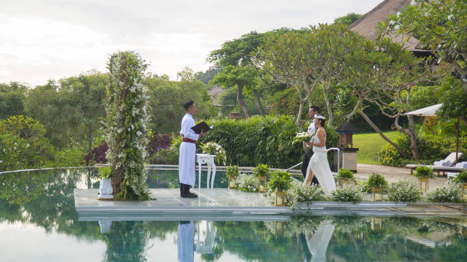 Bride and groom walk across aisle over outdoor pool towards wedding officiant