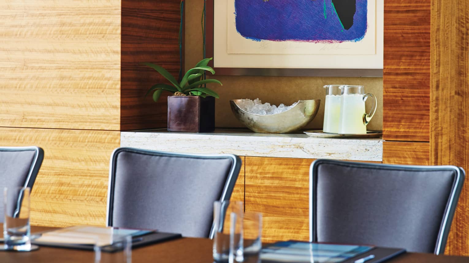 Boardroom table and black meeting chairs in front of modern wood wall, shelf with plant and jugs