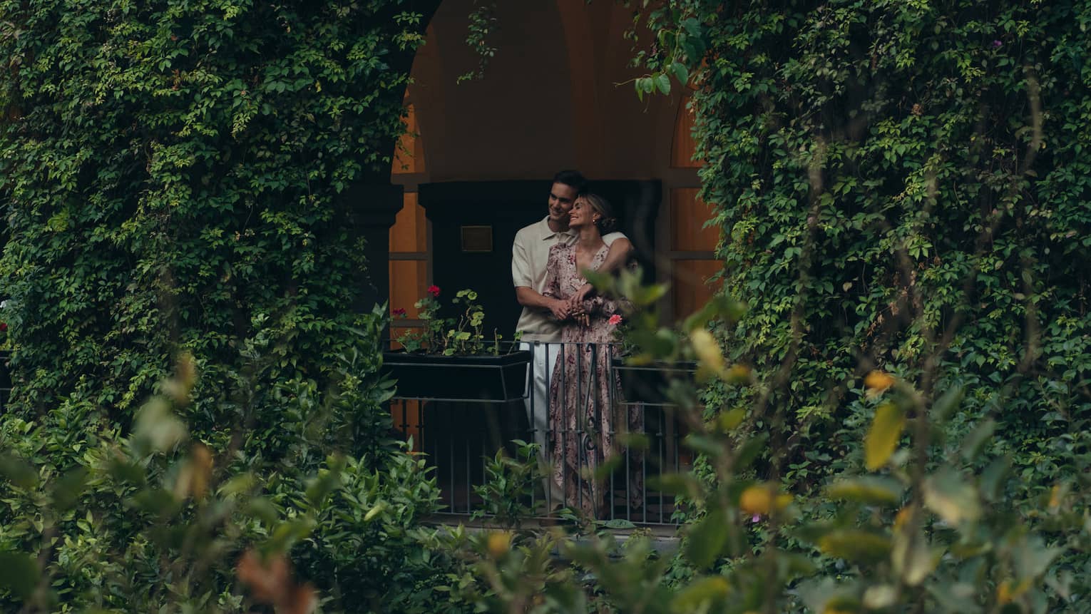 Two people look out on the greenery from behind a railing in an arched opening in a foliage-covered wall