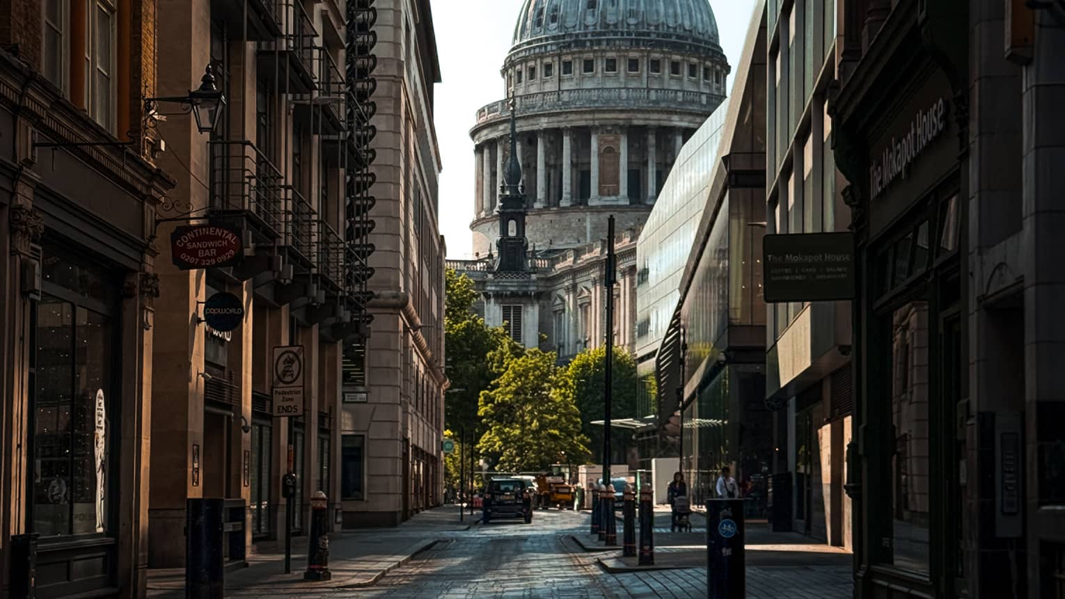 A city street in shadow, lined with buildings. The dome and spire of a cathedral rise in the background, beyond the end of the street.