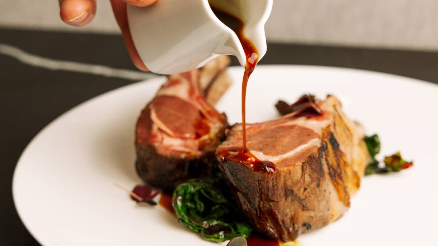 A hand drizzles sauce from a small ceramic saucer onto a rack of lamb, garnished with greens, on a round white plate.